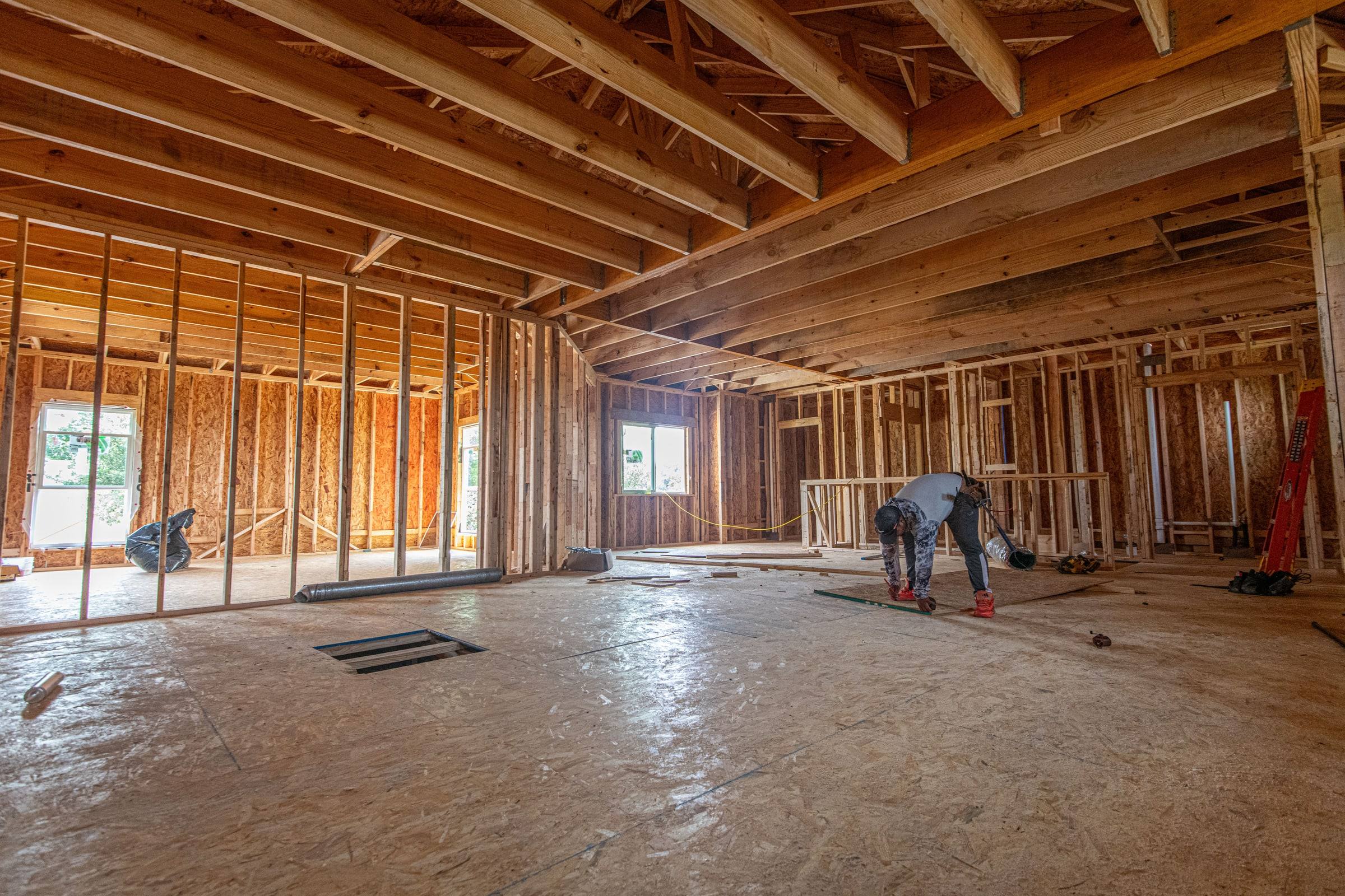 The framed interior of a large new room under construction, open stud walls, ceiling joists and a plywood subfloor, a carpenter at work