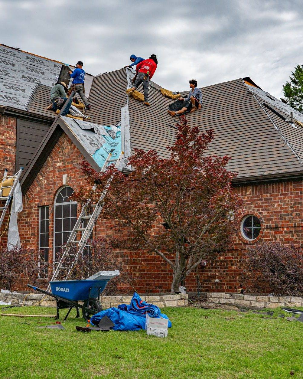 A crew mid tear-off on a brick house, ladders up and the roof stripped back in sections