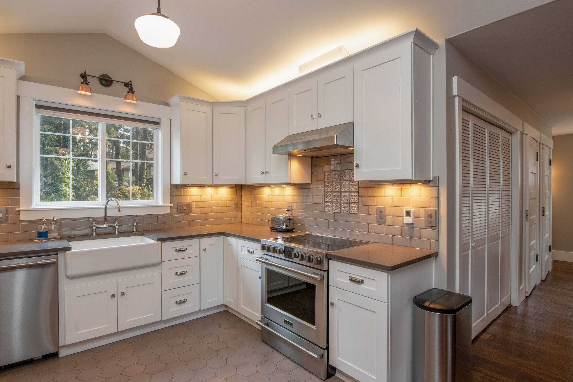 A white kitchen with a farmhouse sink under the window, a stainless range and hood, and a tiled backsplash with outlets