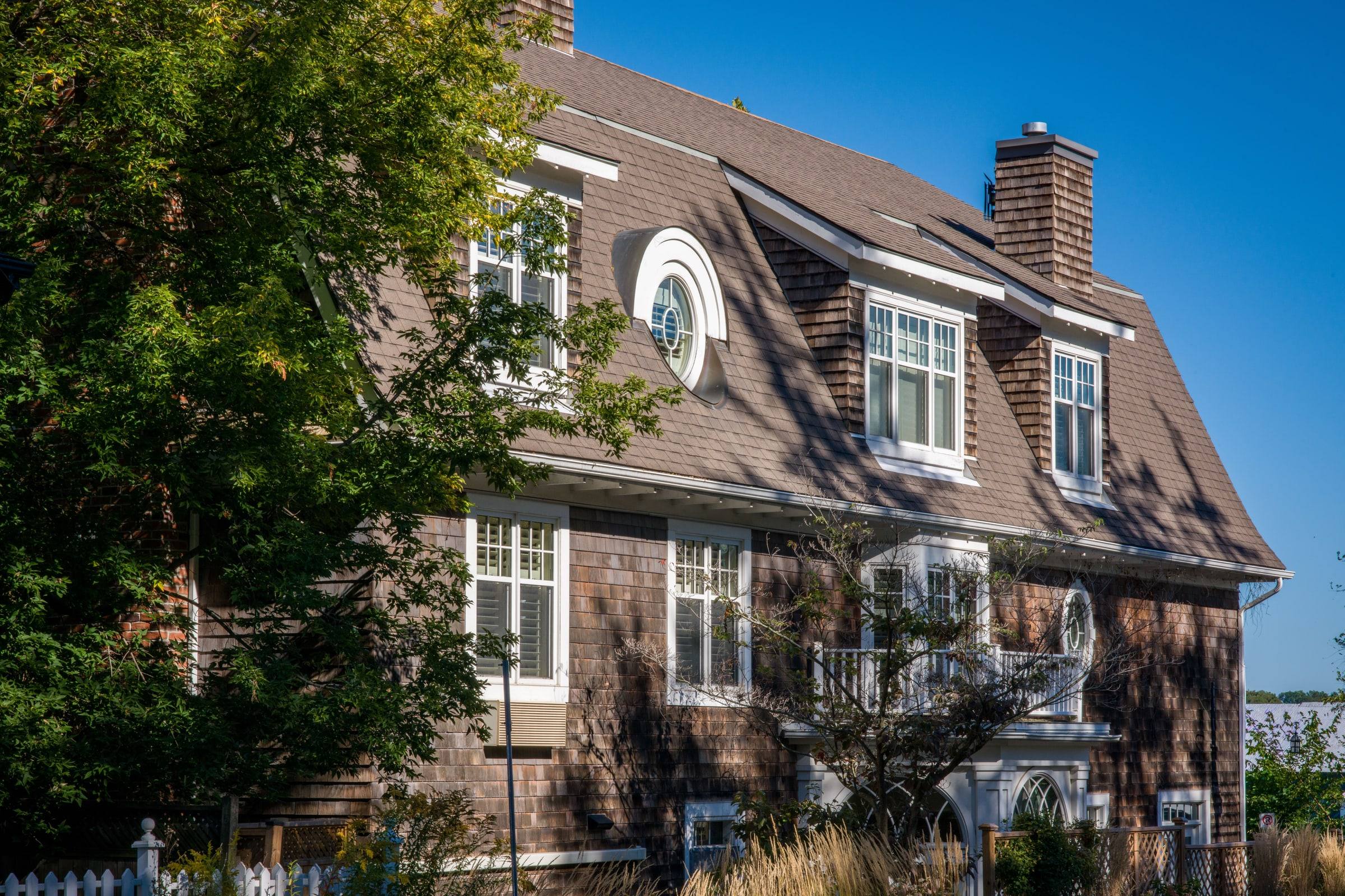 A shingle-sided gambrel house with a large architectural shingle roof and white trim under a clear sky